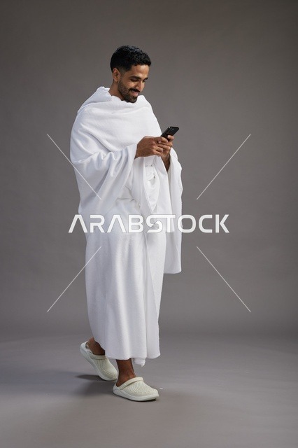 بورتريه لرجل عربي سعودي خليجي يرتدي ملابس الإحرام يستخدم الهاتف المحمول, مفهوم الحج والعمره , كامل الجسم, خلفيه رماديه