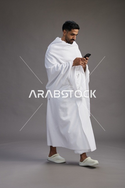 بورتريه لرجل عربي سعودي خليجي يرتدي ملابس الإحرام يستخدم الهاتف المحمول, مفهوم الحج والعمره , كامل الجسم, خلفيه رماديه