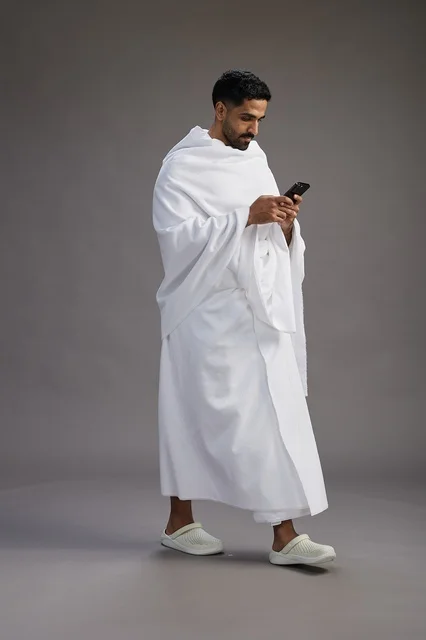 بورتريه لرجل عربي سعودي خليجي يرتدي ملابس الإحرام يستخدم الهاتف المحمول, مفهوم الحج والعمره , كامل الجسم, خلفيه رماديه