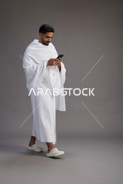 بورتريه لرجل عربي سعودي خليجي يرتدي ملابس الإحرام يستخدم الهاتف المحمول, مفهوم الحج والعمره , كامل الجسم, خلفيه رماديه