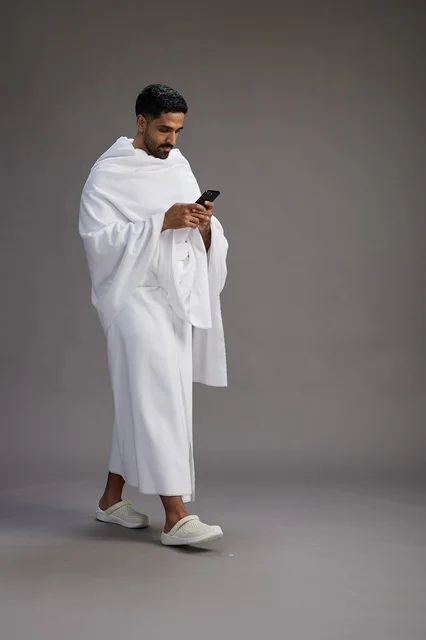 بورتريه لرجل عربي سعودي خليجي يرتدي ملابس الإحرام يستخدم الهاتف المحمول, مفهوم الحج والعمره , كامل الجسم, خلفيه رماديه