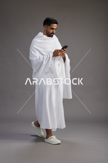 بورتريه لرجل عربي سعودي خليجي يرتدي ملابس الإحرام يستخدم الهاتف المحمول, مفهوم الحج والعمره , كامل الجسم, خلفيه رماديه