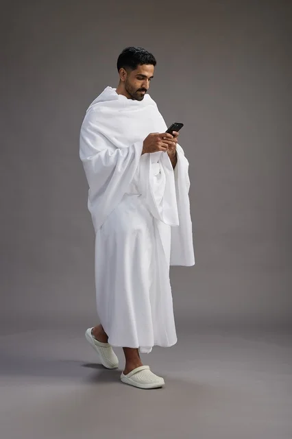 A portrait of an Arab Saudi Gulf man wearing Ihram clothing using a mobile phone, concept of Hajj and Umrah, full body, gray background.