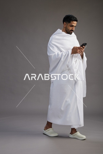 بورتريه لرجل عربي سعودي خليجي يرتدي ملابس الإحرام يستخدم الهاتف المحمول, مفهوم الحج والعمره , كامل الجسم, خلفيه رماديه