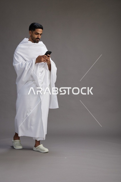 بورتريه لرجل عربي سعودي خليجي يرتدي ملابس الإحرام يستخدم الهاتف المحمول, مفهوم الحج والعمره , كامل الجسم, خلفيه رماديه