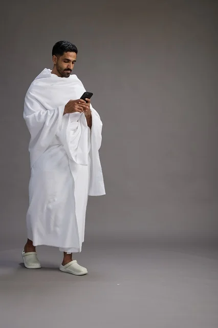 A portrait of an Arab Saudi Gulf man wearing Ihram clothing using a mobile phone, concept of Hajj and Umrah, full body, gray background.