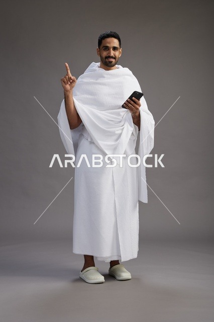 بورتريه لرجل عربي سعودي خليجي يرتدي ملابس الإحرام يستخدم الهاتف المحمول, مفهوم الحج والعمره , كامل الجسم, خلفيه رماديه