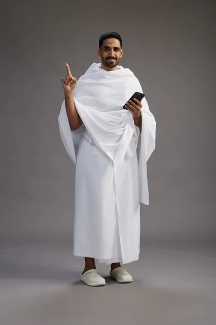 A portrait of an Arab Saudi Gulf man wearing Ihram clothing using a mobile phone, concept of Hajj and Umrah, full body, gray background.