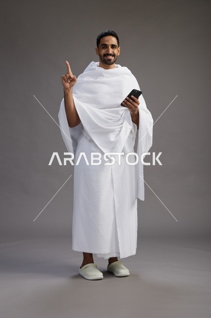 بورتريه لرجل عربي سعودي خليجي يرتدي ملابس الإحرام يستخدم الهاتف المحمول, مفهوم الحج والعمره , كامل الجسم, خلفيه رماديه