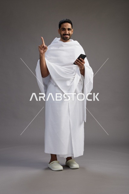 بورتريه لرجل عربي سعودي خليجي يرتدي ملابس الإحرام يستخدم الهاتف المحمول, مفهوم الحج والعمره , كامل الجسم, خلفيه رماديه