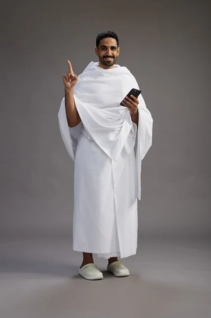 بورتريه لرجل عربي سعودي خليجي يرتدي ملابس الإحرام يستخدم الهاتف المحمول, مفهوم الحج والعمره , كامل الجسم, خلفيه رماديه