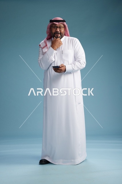بورتريه لرجل  عربي سعودي  خليجي يرتدي الثوب الابيض والشماغ يستخدم الهاتف الذكي, ايماءات وحركات متنوعه , الجسم بالطول الكامل, خلفيه زرقاء
