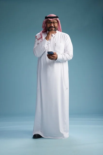A portrait of a Saudi Arabian Gulf man wearing a white thobe and a shemagh using a smartphone, with various gestures and movements, full-body, blue background.