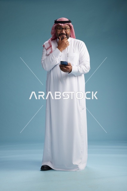 بورتريه لرجل  عربي سعودي  خليجي يرتدي الثوب الابيض والشماغ يستخدم الهاتف الذكي, ايماءات وحركات متنوعه , الجسم بالطول الكامل, خلفيه زرقاء