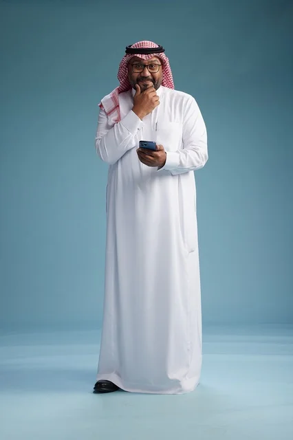 A portrait of a Saudi Arabian Gulf man wearing a white thobe and a shemagh using a smartphone, with various gestures and movements, full-body, blue background.