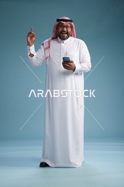 بورتريه لرجل  عربي سعودي  خليجي يرتدي الثوب الابيض والشماغ يستخدم الهاتف الذكي, ايماءات وحركات متنوعه , الجسم بالطول الكامل, خلفيه زرقاء