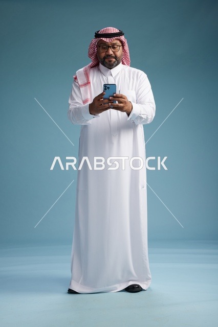 بورتريه لرجل  عربي سعودي  خليجي يرتدي الثوب الابيض والشماغ يستخدم الهاتف الذكي, ايماءات وحركات متنوعه , الجسم بالطول الكامل, خلفيه زرقاء