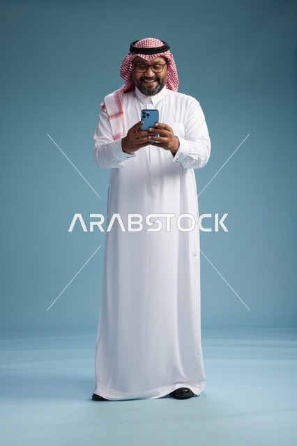 بورتريه لرجل  عربي سعودي  خليجي يرتدي الثوب الابيض والشماغ يستخدم الهاتف الذكي, ايماءات وحركات متنوعه , الجسم بالطول الكامل, خلفيه زرقاء