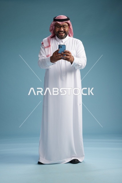 بورتريه لرجل  عربي سعودي  خليجي يرتدي الثوب الابيض والشماغ يستخدم الهاتف الذكي, ايماءات وحركات متنوعه , الجسم بالطول الكامل, خلفيه زرقاء
