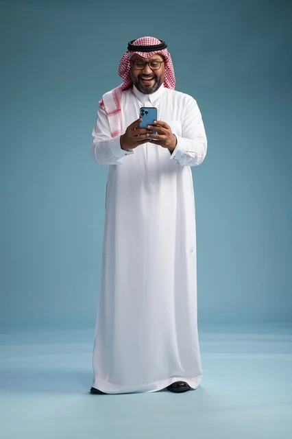 بورتريه لرجل  عربي سعودي  خليجي يرتدي الثوب الابيض والشماغ يستخدم الهاتف الذكي, ايماءات وحركات متنوعه , الجسم بالطول الكامل, خلفيه زرقاء