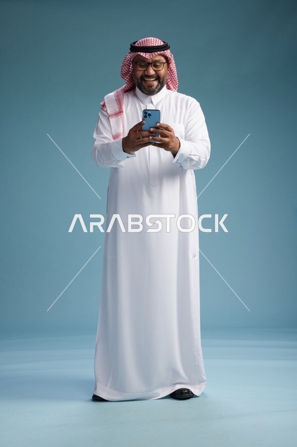 بورتريه لرجل  عربي سعودي  خليجي يرتدي الثوب الابيض والشماغ يستخدم الهاتف الذكي, ايماءات وحركات متنوعه , الجسم بالطول الكامل, خلفيه زرقاء