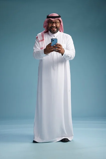 A portrait of a Saudi Arabian Gulf man wearing a white thobe and a shemagh using a smartphone, with various gestures and movements, full-body, blue background. A portrait of a Saudi Arabian Gulf man wearing a white thobe and a shemagh using a smartphone, with various gestures and movements, full-body, blue background.