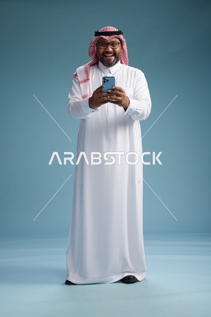 بورتريه لرجل  عربي سعودي  خليجي يرتدي الثوب الابيض والشماغ يستخدم الهاتف الذكي, ايماءات وحركات متنوعه , الجسم بالطول الكامل, خلفيه زرقاء