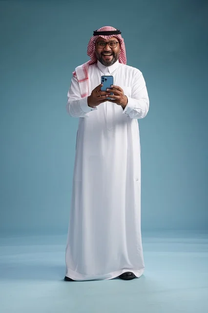 A portrait of a Saudi Arabian Gulf man wearing a white thobe and a shemagh using a smartphone, with various gestures and movements, full-body, blue background.