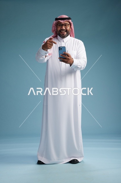 بورتريه لرجل  عربي سعودي  خليجي يرتدي الثوب الابيض والشماغ يستخدم الهاتف الذكي, ايماءات وحركات متنوعه , الجسم بالطول الكامل, خلفيه زرقاء