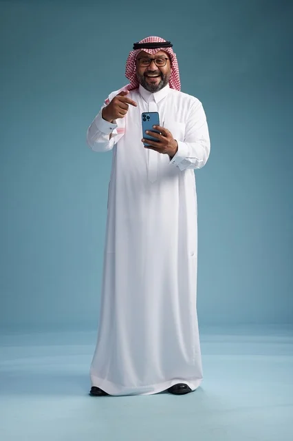 A portrait of a Saudi Arabian Gulf man wearing a white thobe and a shemagh using a smartphone, with various gestures and movements, full-body, blue background.