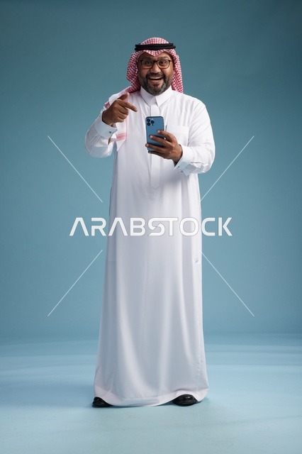 بورتريه لرجل  عربي سعودي  خليجي يرتدي الثوب الابيض والشماغ يستخدم الهاتف الذكي, ايماءات وحركات متنوعه , الجسم بالطول الكامل, خلفيه زرقاء