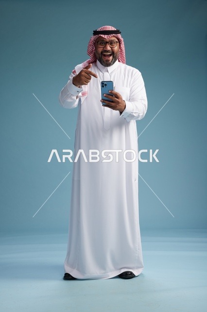 بورتريه لرجل  عربي سعودي  خليجي يرتدي الثوب الابيض والشماغ يستخدم الهاتف الذكي, ايماءات وحركات متنوعه , الجسم بالطول الكامل, خلفيه زرقاء