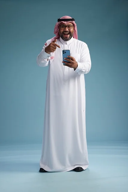 A portrait of a Saudi Arabian Gulf man wearing a white thobe and a shemagh using a smartphone, with various gestures and movements, full-body, blue background.