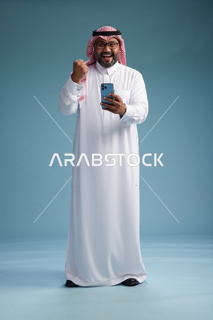 بورتريه لرجل  عربي سعودي  خليجي يرتدي الثوب الابيض والشماغ يستخدم الهاتف الذكي, ايماءات وحركات متنوعه , الجسم بالطول الكامل, خلفيه زرقاء