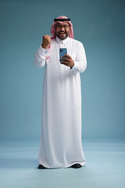 A portrait of a Saudi Arabian Gulf man wearing a white thobe and a shemagh using a smartphone, with various gestures and movements, full-body, blue background.