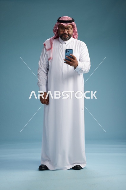 بورتريه لرجل  عربي سعودي  خليجي يرتدي الثوب الابيض والشماغ يستخدم الهاتف الذكي, ايماءات وحركات متنوعه , الجسم بالطول الكامل, خلفيه زرقاء