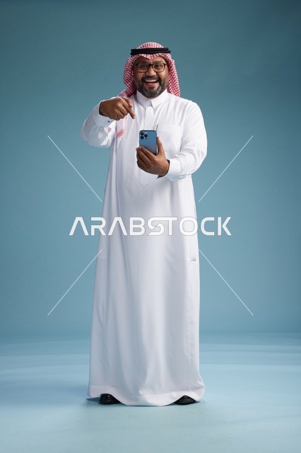 بورتريه لرجل  عربي سعودي  خليجي يرتدي الثوب الابيض والشماغ يستخدم الهاتف الذكي, ايماءات وحركات متنوعه , الجسم بالطول الكامل, خلفيه زرقاء