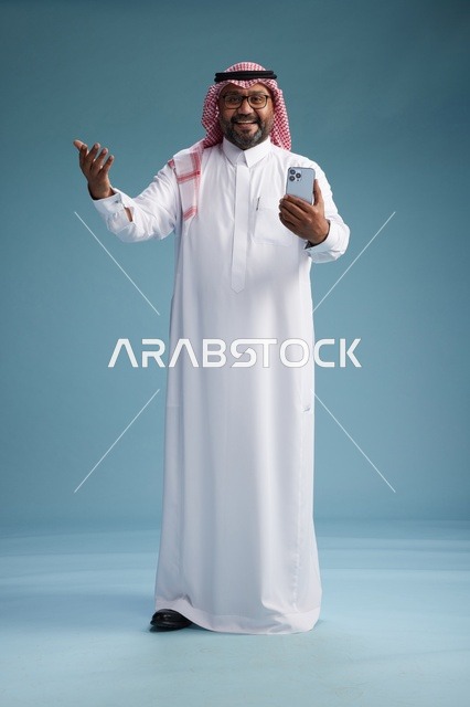 بورتريه لرجل  عربي سعودي  خليجي يرتدي الثوب الابيض والشماغ يستخدم الهاتف الذكي, ايماءات وحركات متنوعه , الجسم بالطول الكامل, خلفيه زرقاء
