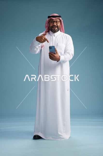 بورتريه لرجل  عربي سعودي  خليجي يرتدي الثوب الابيض والشماغ يستخدم الهاتف الذكي, ايماءات وحركات متنوعه , الجسم بالطول الكامل, خلفيه زرقاء
