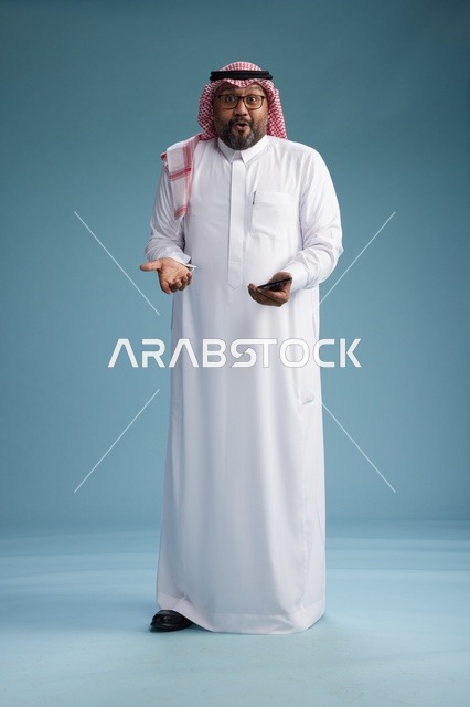 بورتريه لرجل  عربي سعودي  خليجي يرتدي الثوب الابيض والشماغ يستخدم الهاتف الذكي, ايماءات وحركات متنوعه , الجسم بالطول الكامل, خلفيه زرقاء