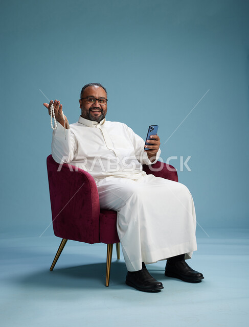 رجل سعودي يرتدي الثوب الابيض يجلس على كرسي احمر يستخدم الهاتف المحمول, حركات وايماءات منوعه, خلفيه زرقاء