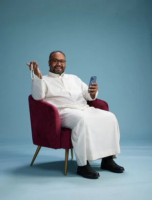 A Saudi man wearing a white thobe sits on a red chair using a mobile phone, with various movements and gestures, against a blue background.