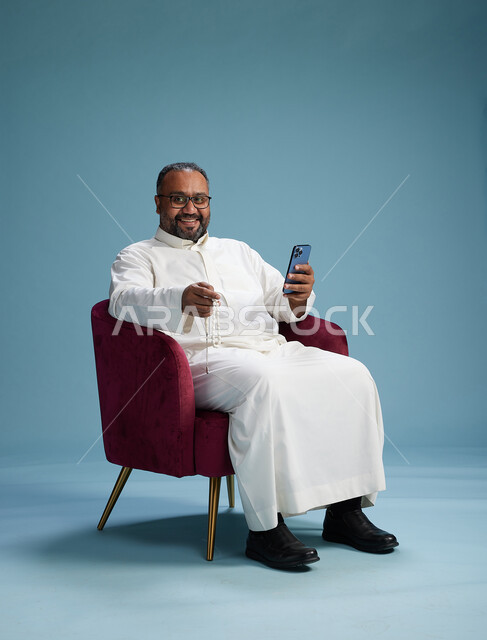 A Saudi man wearing a white thobe sits on a red chair using a mobile phone, with various movements and gestures, against a blue background.