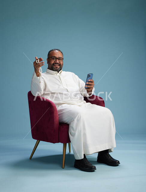 رجل سعودي يرتدي الثوب الابيض يجلس على كرسي احمر يستخدم الهاتف المحمول, حركات وايماءات منوعه, خلفيه زرقاء