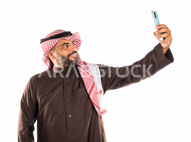 بورتريه لرجل عربي خليجي سعودي يقوم بالتقاط صورة تذكارية من خلال الهاتف المحمول، صور تذكارية، صور سيلفي، كاميرا امامية، مكالمة فيديو، بث فيديو مباشر، ايماءات وجه مبتسمة، خلفية بيضاء