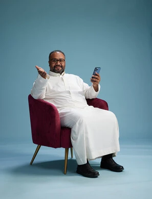 A Saudi man wearing a white thobe sits on a red chair using a mobile phone, with various movements and gestures, against a blue background.