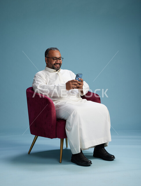 A Saudi man wearing a white thobe sits on a red chair using a mobile phone, with various movements and gestures, against a blue background.