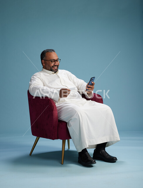 بورتريه لرجل سعودي جالس يستخدم الهاتف المحمول ويحمل بطاقة ائتمان, مفهوم الدفع الالكتروني عبر بطاقة البنك, خلفيه بيضاء