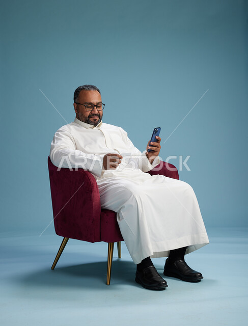 بورتريه لرجل سعودي جالس يستخدم الهاتف المحمول ويحمل بطاقة ائتمان, مفهوم الدفع الالكتروني عبر بطاقة البنك, خلفيه بيضاء