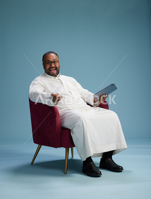 A portrait of a Saudi man using a tablet and a credit card while sitting, the concept of electronic payment via a bank card, blue background.